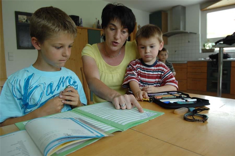 Das Foto zeigt eine Mutter am Tisch mit zwei Kindern, die ein Buch vorliest, und ein weiteres im Hintergrund.