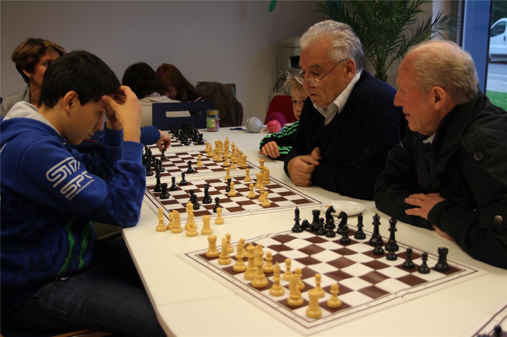 Schachspiel im Jugend- und Stadtteilhaus Schicksbaum innen, auf einem Tisch sind mehrere Schachbretter aufgestellt, zwei ältere Männer spielen gegen Kinder 