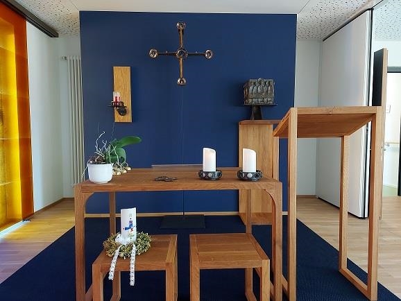 Wir blicken über Bänke zu einem Holzaltar mit blauem Hintergrund und einem Kreuz.
