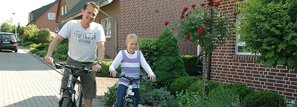 Ein Vater und seine Tochter sitzen auf ihren Fahrrädern mit einem Fuß auf dem Boden vor einem Wohnhaus
