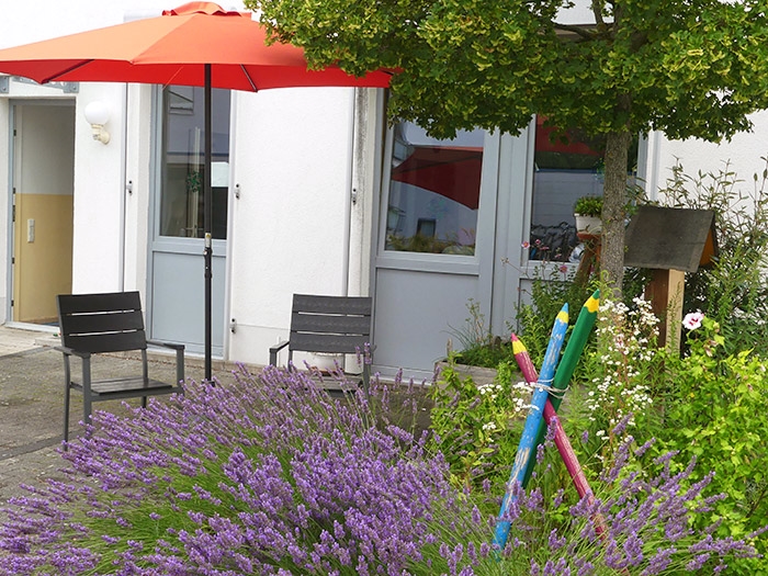 Sitzbereich auf einer Terrasse mit Stühlen und Schirm, davor ein Garten mit Lavendelbusch und eine Skulptur aus drei überdimensionalen Buntstiften (Caritasverband Darmstadt e. V.)