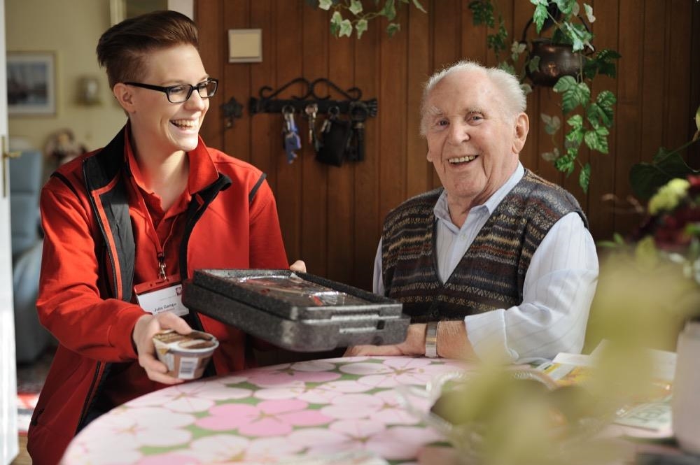 Eine Mitarbeiterin des Menüdienst reicht einem Senior ein Mittagessen 