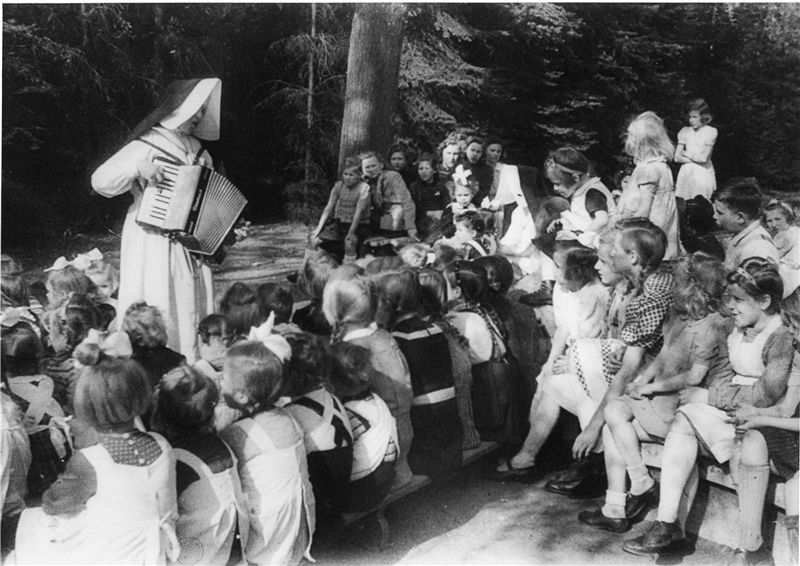 Das Foto zeigt eine Ordensschwester, die einer Schar Kinder vor ihr Akkordeon vorspielt.