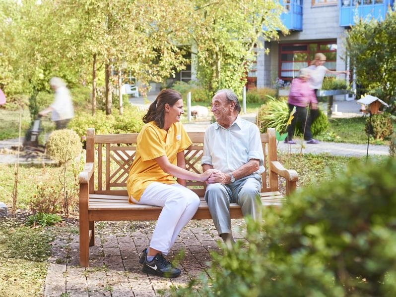Älterer Herr und Pflegerin sitzen auf Holzbank im Garten 