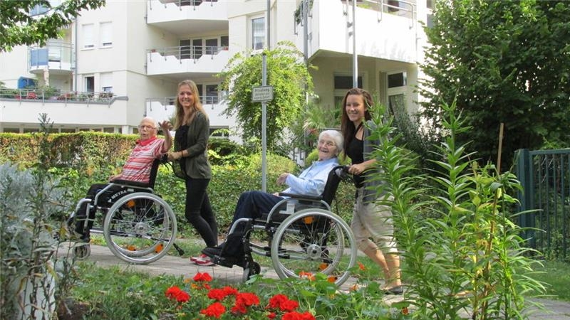 Zwei junge Frauen schieben zwei ältere Damen im Rollstuhl durch den Garten