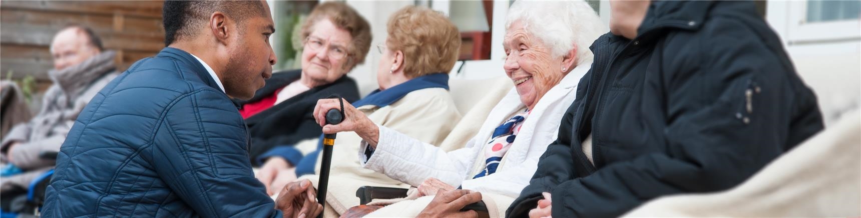Altenpfleger hockt vor sitzender Seniorin und beide blicken sich freundlich an