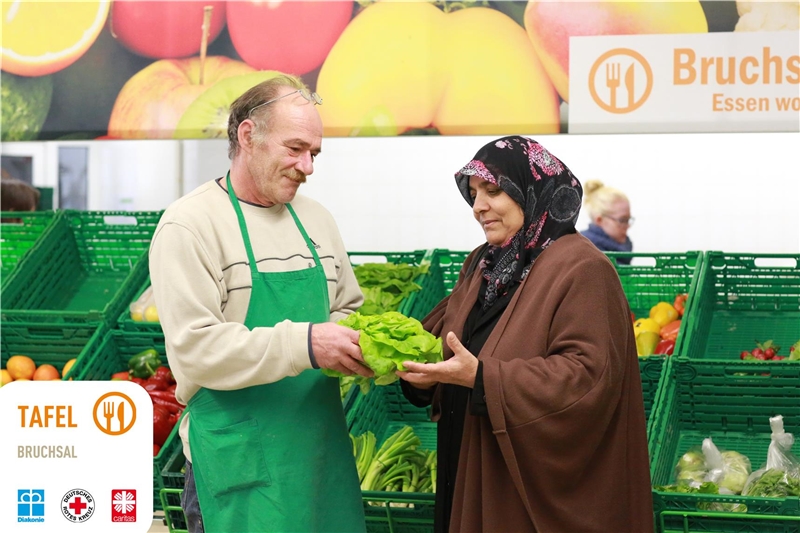 Zwei ältere Menschen stehen in einem Gemüseladen. Der Verkäufer zeigt der Dame einen Kopfsalat. 