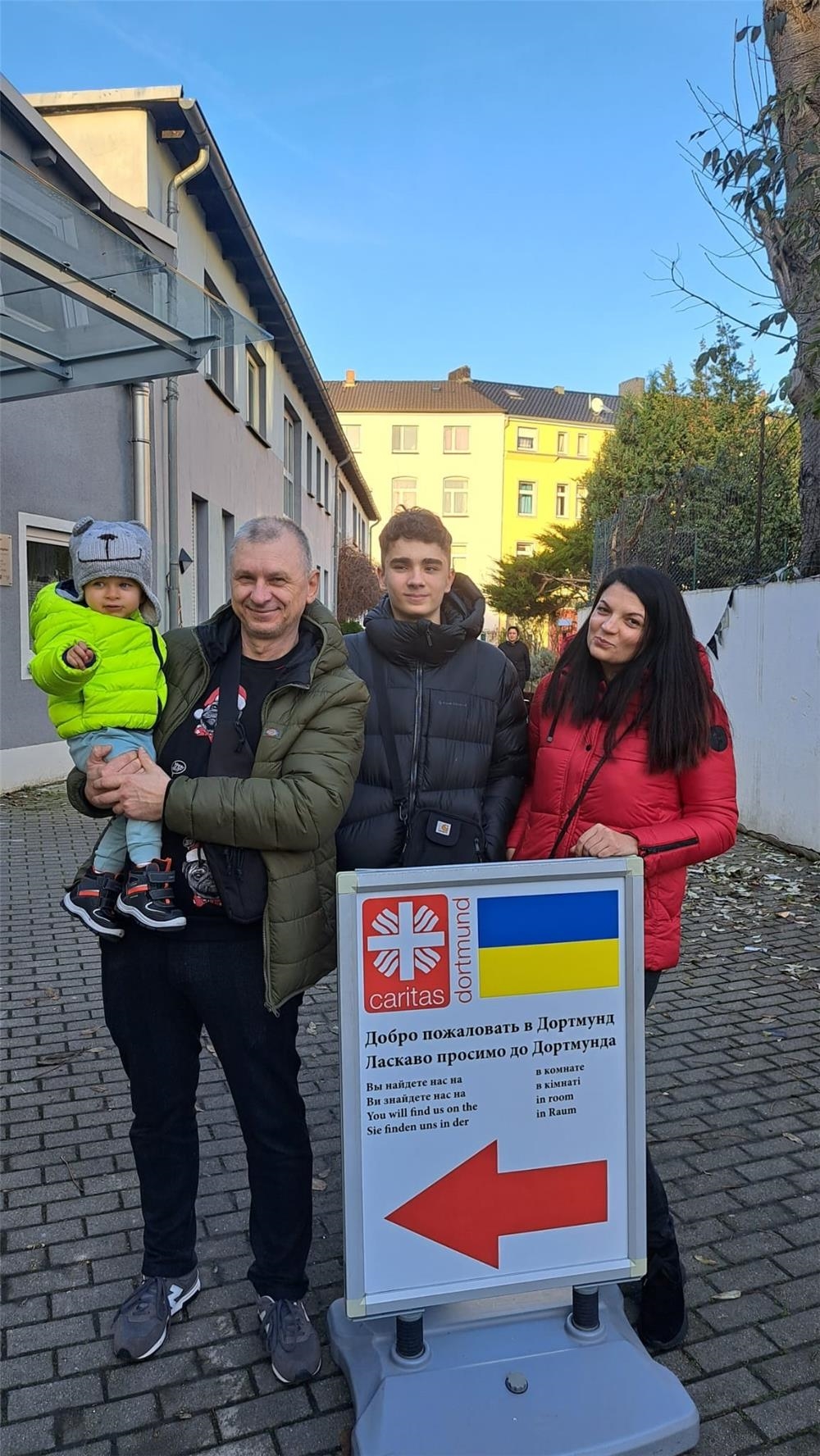 Ein Mann mit einem Kleinkind auf dem Arm, ein Jugendlicher und eine Frau stehen vor dem Wegweiser zur Anblaufstelle der Ukrainehilfe