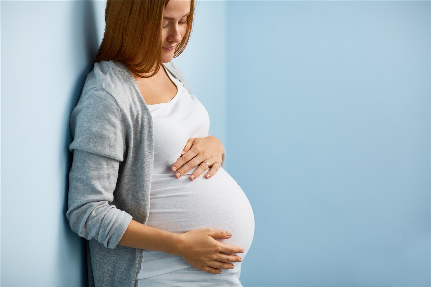 Schwangere Frau schaut auf ihren Bauch vor hellblaue Wänden (AdobeStock_138952178)