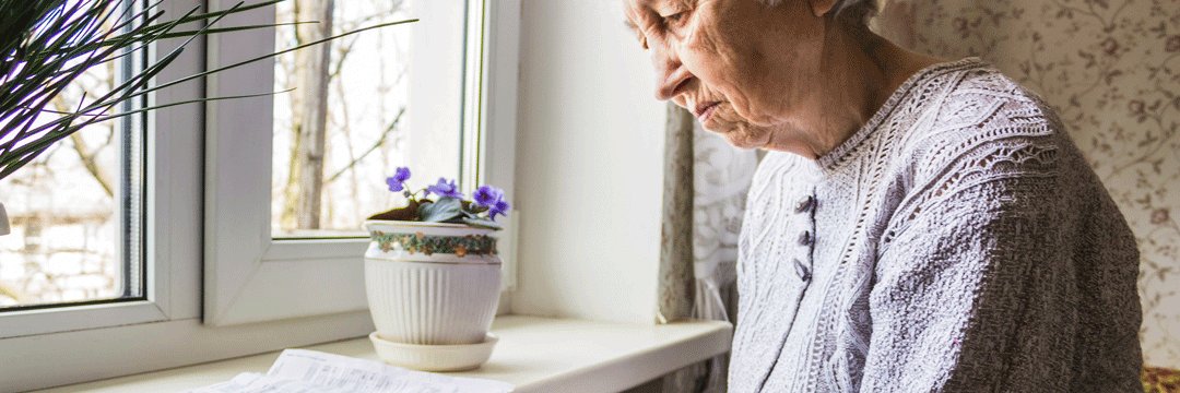 Eine ältere Frau steht am Fenster und blickt traurig auf einen Zettel