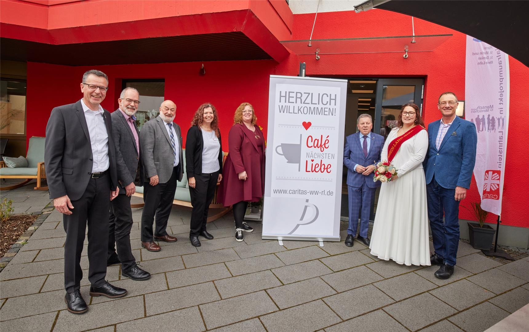Die Caritas-Verantwortlichen und Ehrengäste stehen um das Schild "Herzlich Willkommen im Café Nächstenliebe". (Caritasverband Westerwald-Rhein-Lahn e.V. / Olaf Nitz)