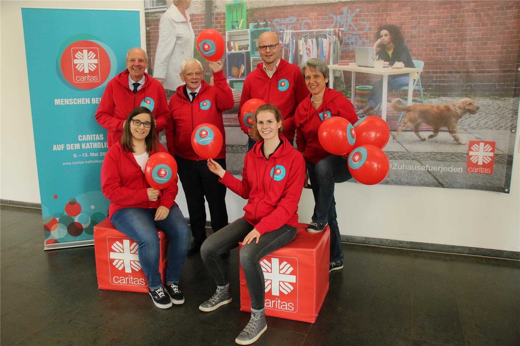 Das 6-köpfige Vorbereitungsteam des Caritasverbandes für die Diözese Münster hat rote Caritas-Jacken an und hält rote Caritas-Luftballons hoch. 