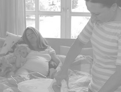 Eine Familienpflegerin steht vor einem Bügelbrett und bügelt ein kleines T-Shirt. Im Hintergrund liegt eine Schwangere mit ihrem Kind auf einem Sofa. Die Aufnahme ist in schwarz-weiß.