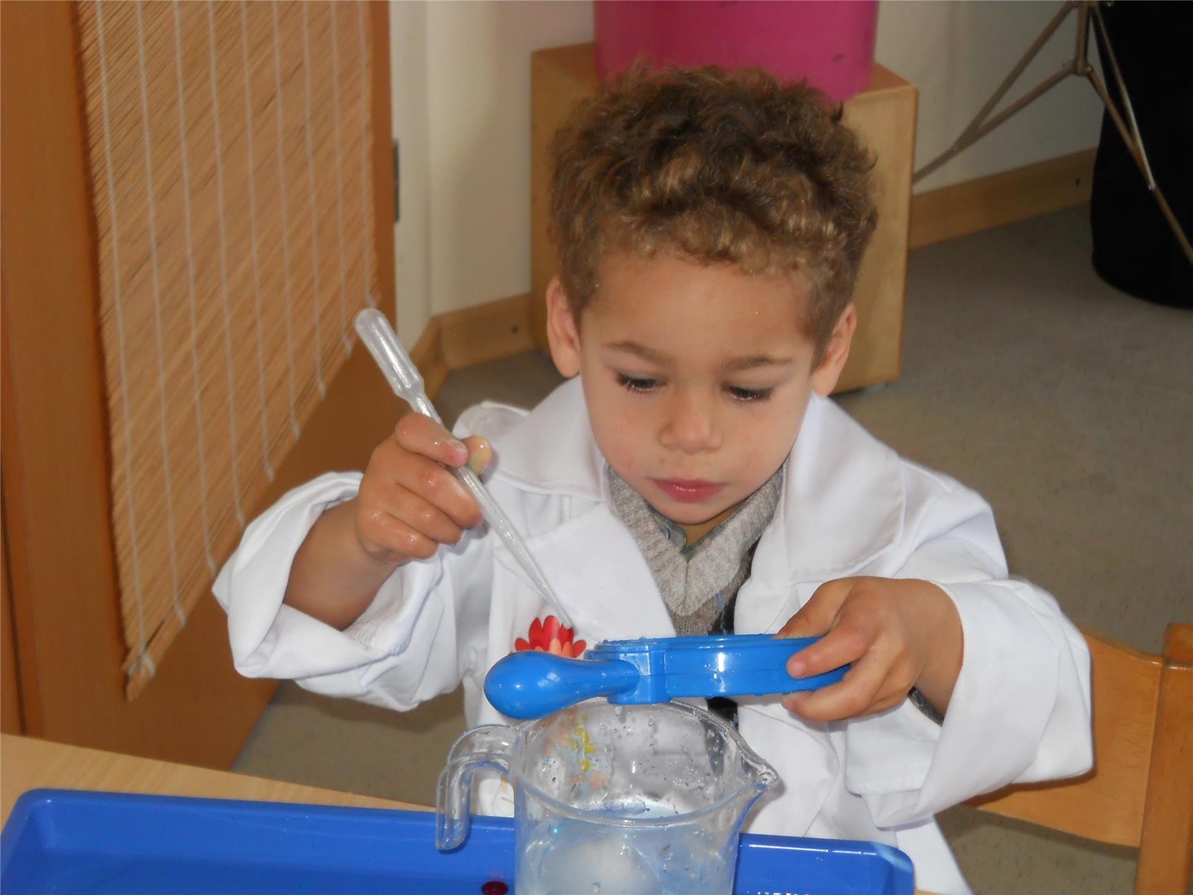 Ein Kind in weißem Kittel sitzt mit einer Pipette und einer Plastikschale in Händen vor einem Tisch, auf dem ein Messbecher mit Wasser steht ( Caritasverband Trier e. V.)