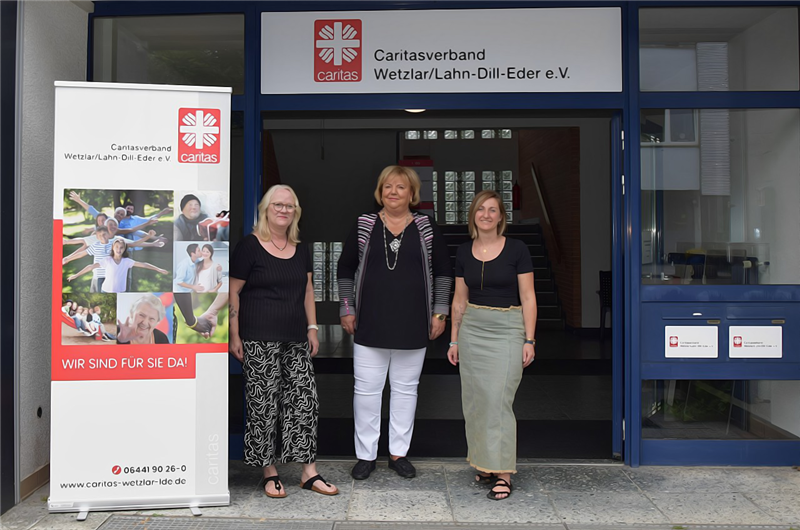 Drei Frauen stehen lächelnd vor dem Eingang des Gebäudes des Caritasverbands Wetzlar/Lahn-Dill-Eder e.V. Links daneben ist ein Banner mit dem Caritas-Logo zu sehen.