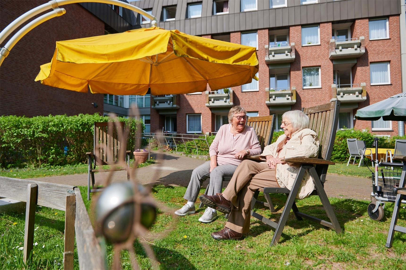 Zwei Frauen sitzen auf Holzstühlen in einem sonnigen Garten 