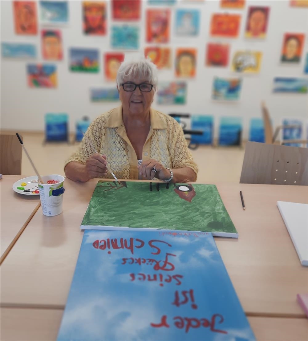 Eine Seniorin sitzt mit Pinsel an einem Tisch und malt ein Bild 