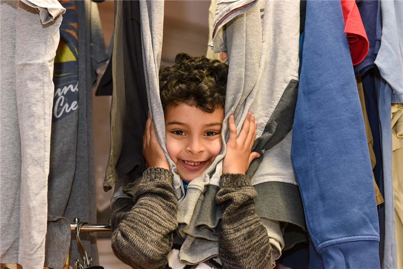 Portrait eines kleinen Jungen, der sein Gesicht zwischen den Klamotten eines Kleiderständers hervorstreckt und lacht