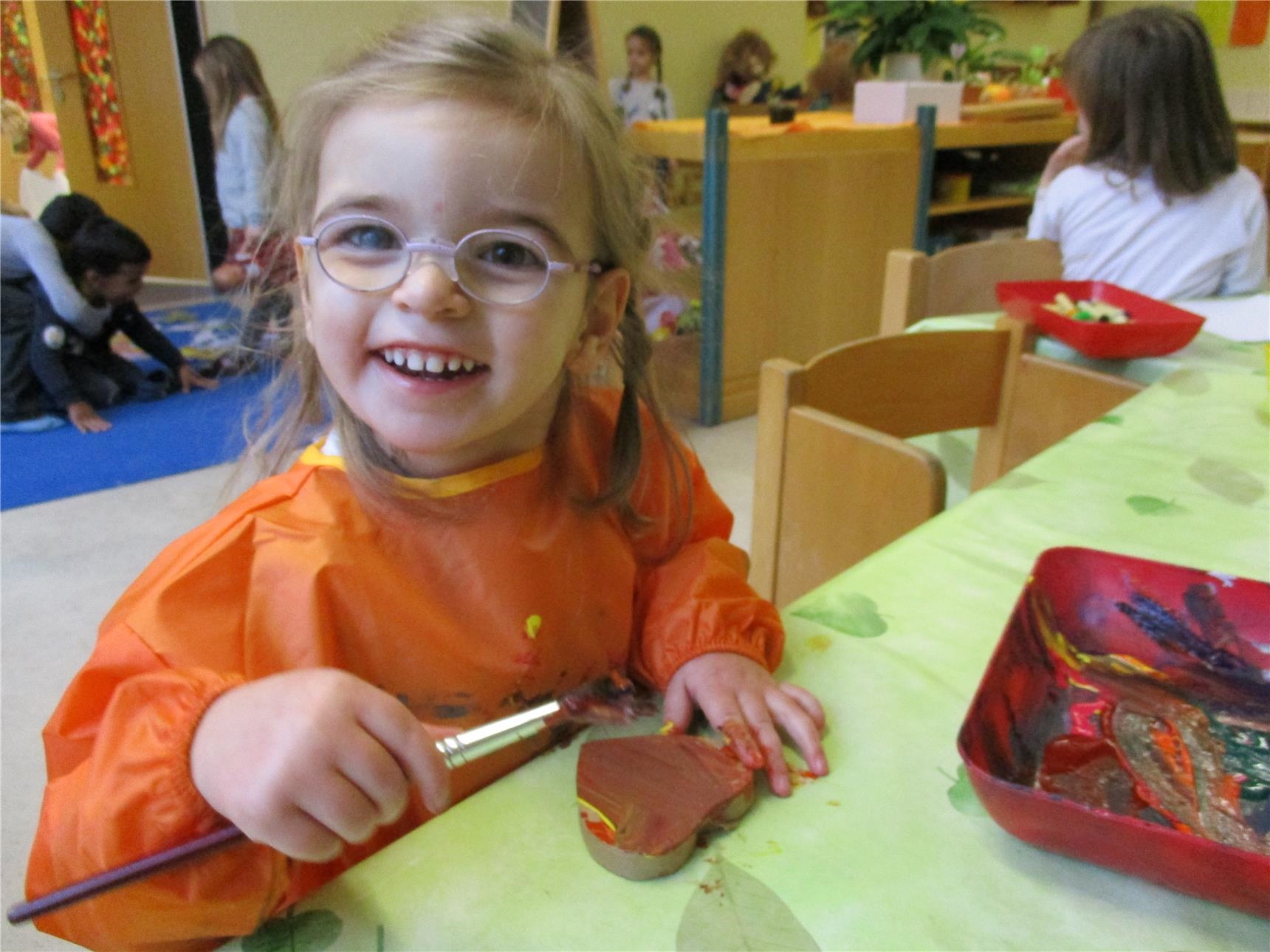 : Ein Mädchen bemalt ein Pappherz mit Pinsel und Farbe; im Hintergrund: spielende Kinder  ( Caritasverband Trier e. V.)