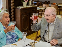 Eine ältere Dame und ein Herr sitzen an einem festlich gedeckten Tisch und prosten sich zu. / Werner Krüper