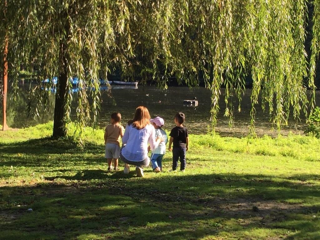 Frau kniet neben Kindern unter Bäumen und schaut auf einen See. 
