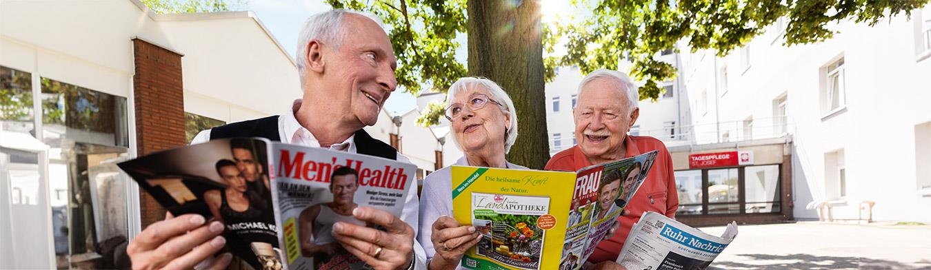 Drei Senior:innen sitzen auf einer Bank und lesen Zeitschriften.