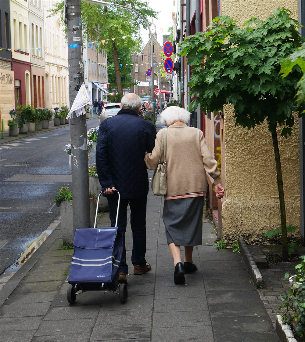 Ein altes Ehepaar läuft den Bürgersteig der Körnerstraße in Köln-Ehrenfeld entlang, der Mann zieht dabei eine blaue Einkaufskarre hinter sich her. Das Ehepaar wurde von hinten fotografiert. (Julia Hitschfeld/Caritas)
