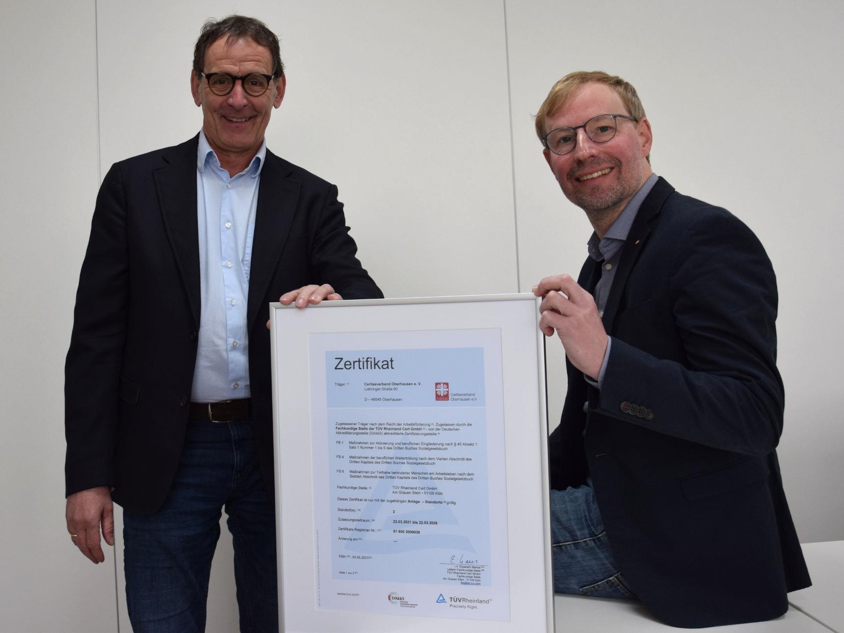 Jürgen Monz und Michaelkreuzfelder halten einen Bilderrahmen mit dem AZAV-Zertifikat der Caritas Oberhausen.
