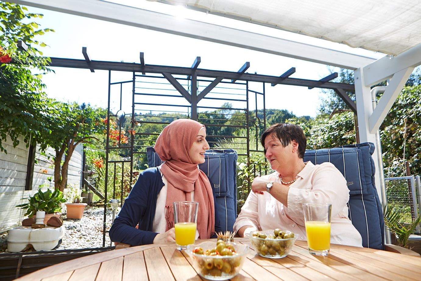 Zwei Frauen sitzen am Tisch und unterhalten sich, eine trägt ein Kopftuch.