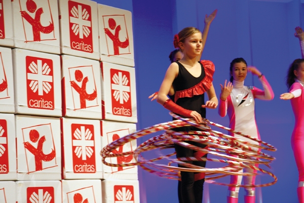 Ein Mädchen während der Bühnenshow zum Auftakt von youngcaritas im Kostüm, die mit rot-weißen Hula Hoop-Reifen spielt. Im Hintergrund steht eine Wand aus Sitzwürfeln mit Caritas-Logos u. zwei Mädchen. (Caritas)