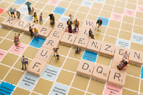 Ein Scrabble-Spielbrett, auf dem die Wörter Kompetenzen, Beruf und DQR liegen. Auch stehen auf dem Brett einige kleine Modellfiguren..