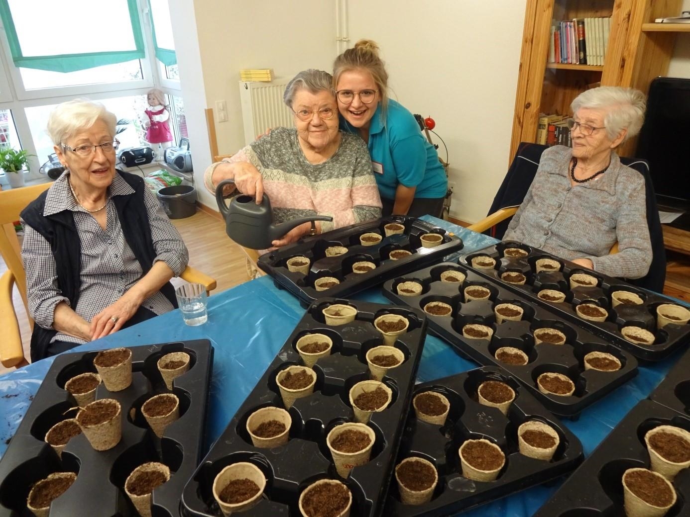 Drei Seniorinnen sitzen am Tisch, auf dem sieben Paletten bestückt mit Erde gefüllten Blumentöpfen, eine Mitarbeiterin steht lachend hinter einer Seniorin (Seniorenzentrum St. Bonifatius)
