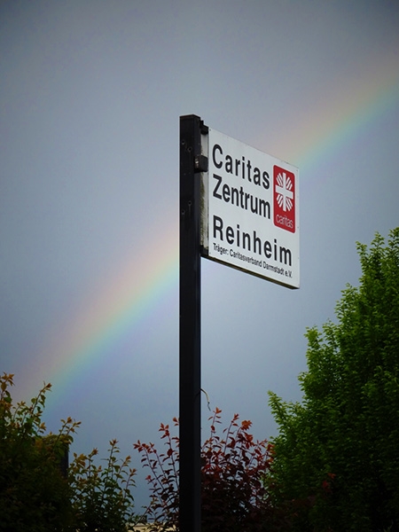 Wegweiser-Schild zum Caritas Zentrum Reinheim mit Regenbogen dahinter (Caritasverband Darmstadt e. V.)