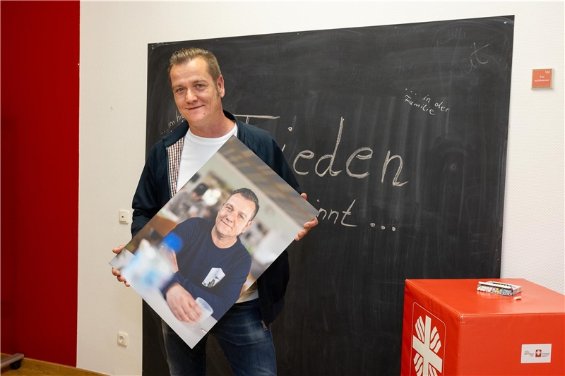 Ein Mann hält ein Fotoportrait von sich in die Kamera vor einer Tafel mit der Aufschrift "Frieden beginnt"