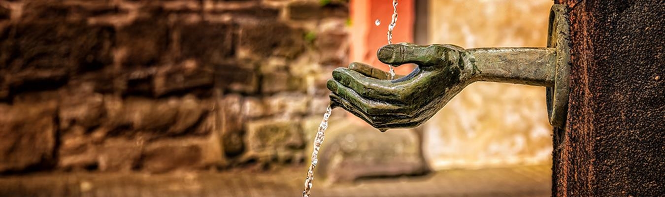 Bronzene Hand-Skulptur mit fließendem Wasser vor einer rustikalen Steinmauer, symbolisch für Großzügigkeit und Fürsorge.