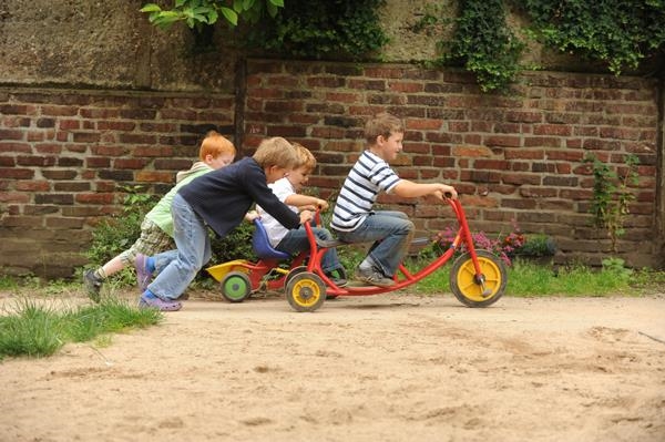drei Kinder schieben einen Junge auf einem Dreirad