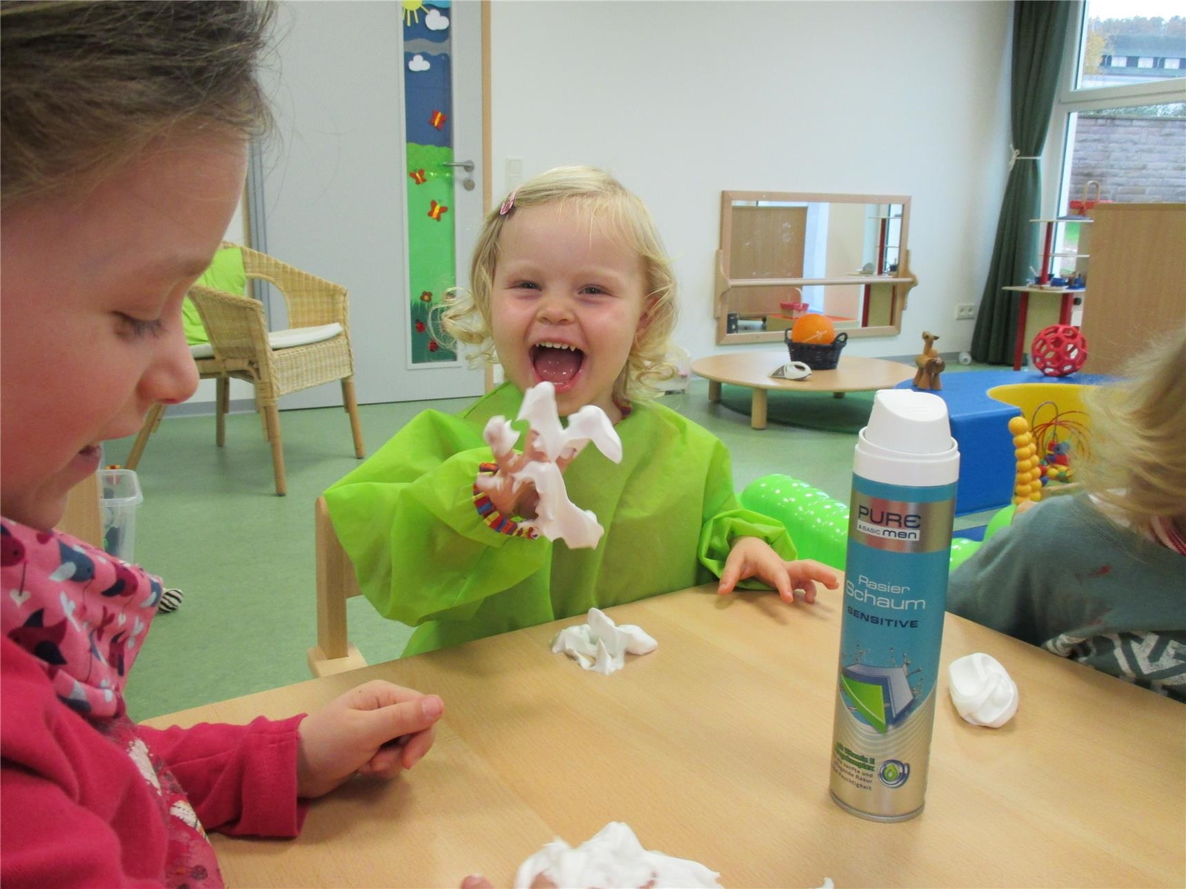 Auf einem Tisch befinden sich eine Dose Rasierschaum und drei Kleckse Rasierschaum; ein Kind hat die Finger voll Rasierschaum und lacht (Caritasverband Trier e. V.)
