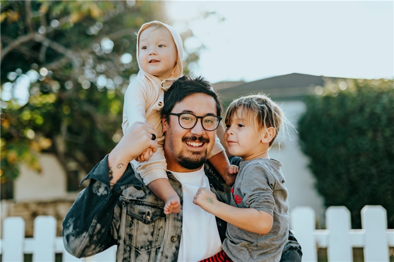 Ein dunkelhaariger Mann mit Brille und Bart lächelt in die Kamera und hält ein Kleinkind auf dem Arm und ein Baby auf den Schultern