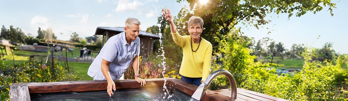 Eine Seniorin und eine Pflegerin bespritzen sich an einem Brunnen mit Wasser.