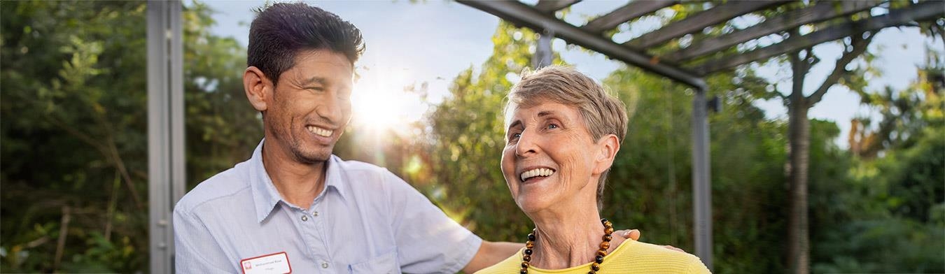 Ein Pfleger und eine Seniorin stehen im Freien und lachen gemeinsam.