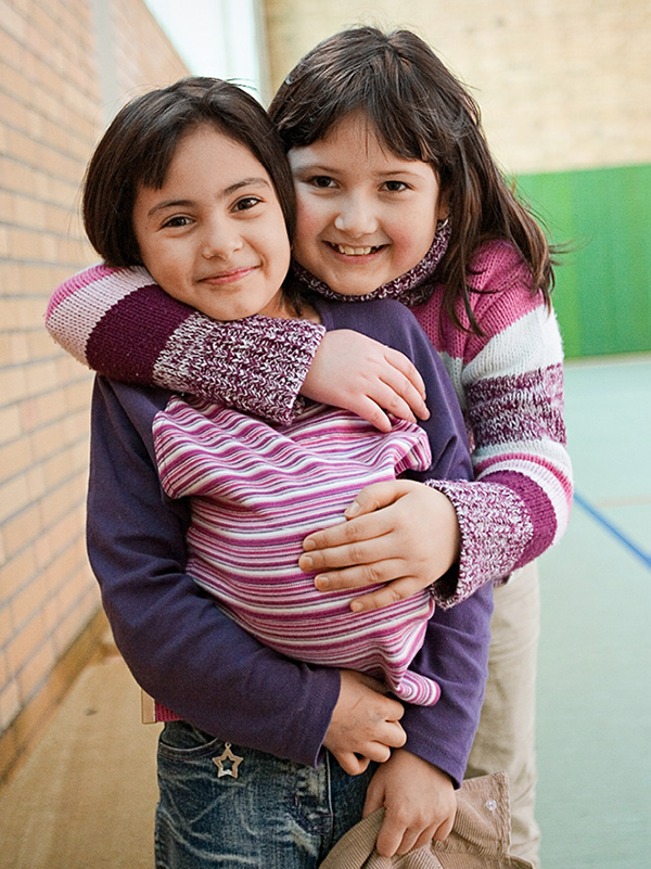 Zwei Mädchen stehen in einer Turnhalle Arm in Arm zusammen und blicken lächelnd in die Kamera. Eines der Mädchen trägt einen Ball unter dem Oberteil. (Foto: Achim Pohl)