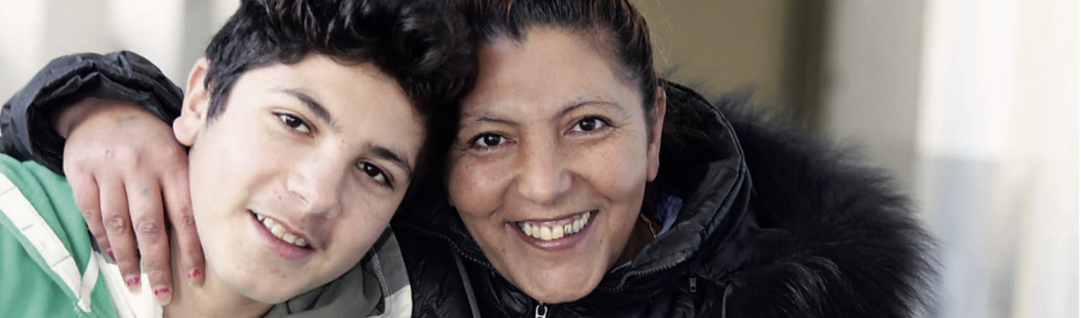 Ein geflüchteter Junge mit seiner Mutter in einer Einrichtung der Caritas. Beide lächeln in die Kamera. 
