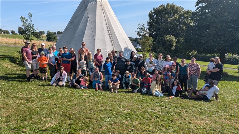 Eine große Gruppe von Menschen unterschiedlichen Alters posiert vor einem Tipi an einem sonnigen Tag auf einer Wiese mit Bäumen im Hintergrund.