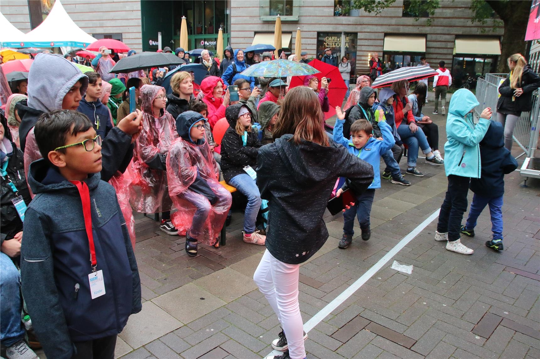 Das Foto zeigt ein geduldig im Regen ausharrendes Publikum und tanzende Kinder davor. (Harald Westbeld)