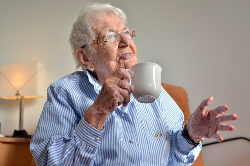 Seniorin sitzt am Tisch und hält Kaffeetasse hoch (Tobias Schneider, taylormade)