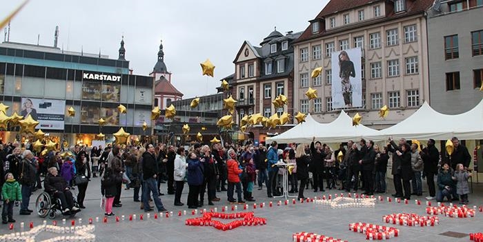 Wir befinden uns auf dem Uniplatz in Fulda. Viele goldene Luftballos im Sternenformat werden von Passanten gehalten. Im Vordergrund sehen wir einen Teil der Kerzen, welche zur diesjährigen Aktion scho