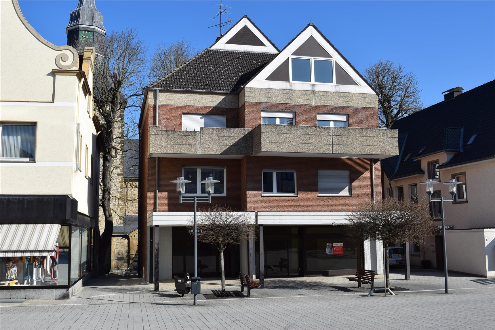 Vorderansicht ein mehrstöckiges braunes Backsteingebäude mit Spitzdach und Balkonen. Im Erdgeschoß liegt die Tagespflege mit großer Glasfront und schützendem Vorbau in weiß direkt und ebenerdig mit dem Marktplatz